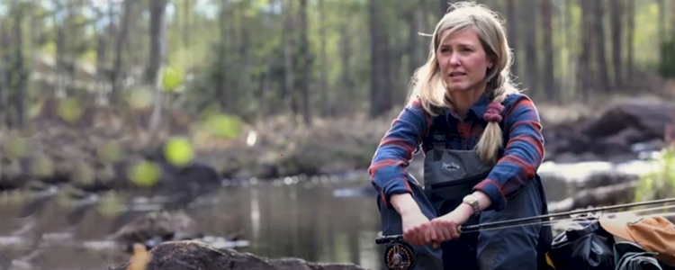 A woman sits by a stream holding a fishing rod. The image is from the film Not Just Fish. Foto: Bitzer Productions AB