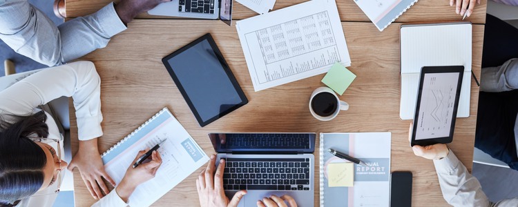 Problemlösning och planering runt en skrivbord på ett kontor. Fotot är taget uppifrån så att man anar fem personer runt bordet, datorer, anteckningsmaterial, postit-lappar, Ipads, kaffekopp med mera. Foto: Mostphotos