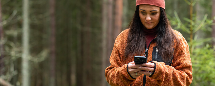 Medelålders kvinna i mössa och fleecejacka tittar på sin mobiltelefon i skogen. Foto: Bulldozer
