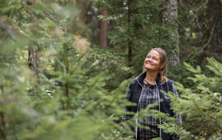 Kvinna klädd i Skogsstyrelsens väst står och tänker i en granskog, hon tittar leende uppåt.