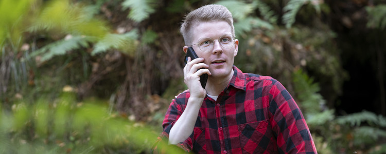 En man i röd och svart rutig skjorta pratar i mobiltelefon i skogen.  Foto: @ Camilla Zilo