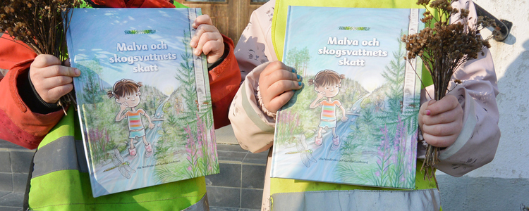Två förskolebarn i ytterkläder håller i varsitt exemplar av barnboken "Malva och Skogsvattnets skatt".