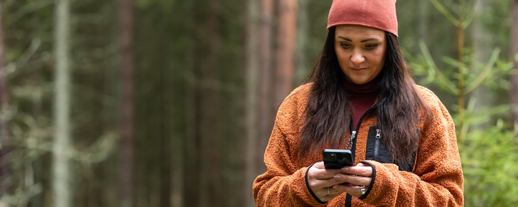 Medelålders kvinna i mössa och fleecejacka tittar på sin mobiltelefon i skogen.