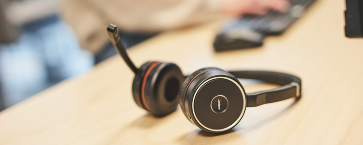 Närbild på headset som ligger på ett skrivbord. I bakgrunden syns någon som skriver på ett tangentbord.