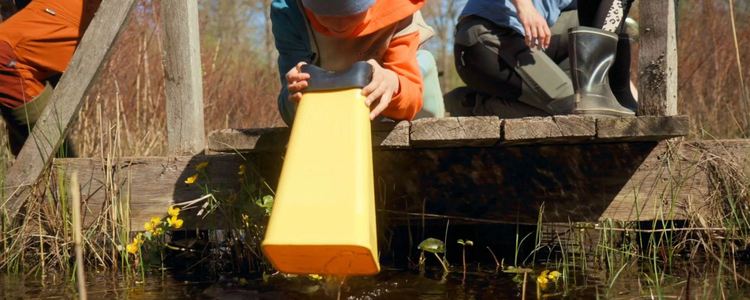 Ett barn knäböjer på en träbrygga och kikar med en stor gul vattenkikare ner i vattnet.