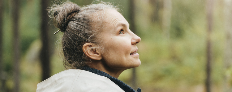 Närbild på en medelålders kvinna iklädd vit regnjacka som går i en tallskog och förnöjsamt tittar upp mot trädkronorna. Bilden är tagen bakifrån så man ser kvinnans axlar och ansiktet syns i profil.  Hon tänker, ser glad och tillfreds ut, som om hon tagit ett smart beslut.