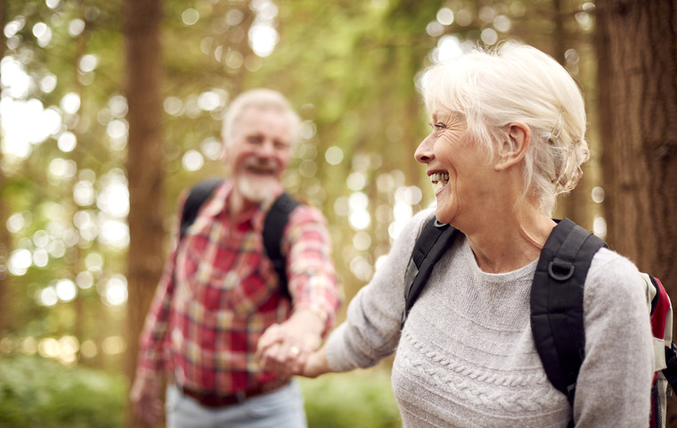 Pensionärer på skogsutflykt. Med varsin ryggsäck på ryggen så skrattar de och håller varandra i handen. Foto: Mostphotos