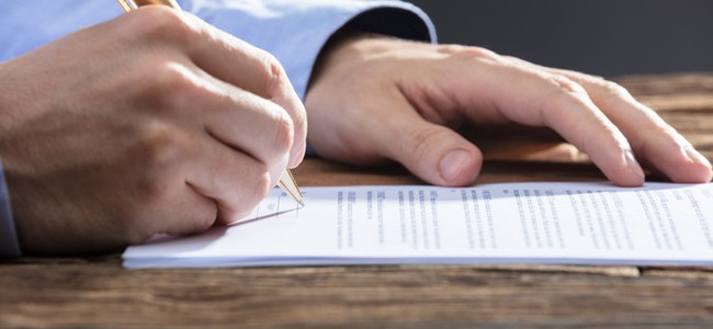 Close-up Of A Businesspersons Hand Signing Document On Wooden Desk