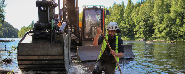 En grävmaskin står i ett större vattendrag i Västerboggen. Restaureringsarbete pågår. I förgrunden, strax framför grävskopan i vattnet, står en man i varselväst och vit hjälm. Mannen guidar maskinföraren. Foto: Linnéa Rådahl, Länsstyrelsen Västerbotten