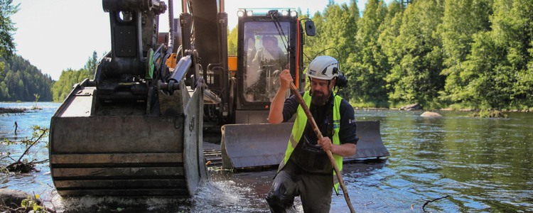 An excavator stands in a larger stream where restoration work is underway. In the foreground, just in front of the excavator’s bucket in the water, stands a man wearing a high-visibility vest and white helmet. Foto: Linnéa Rådahl, Länsstyrelsen Västerbotten