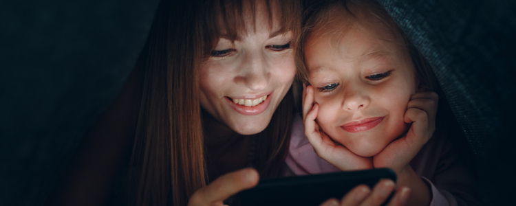 Mor och dotter ligger under en filt i mörkret och tittar på en mobiltelefon.