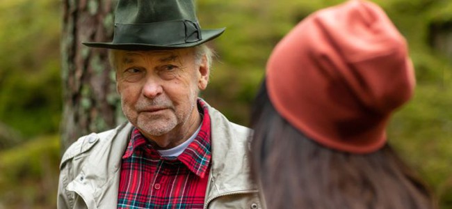 Äldre man i hatt, jacka och rutig skjorta och medelålders kvinna i mössa och fleecejacka pratar med varandra i skog.