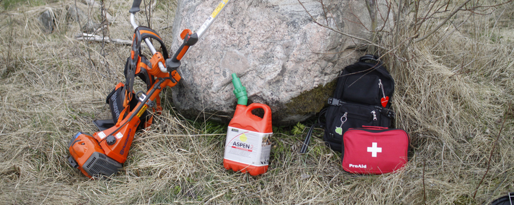 Röjsåg, ryggsäck och första hjälpen-kit står på marken framför en sten.