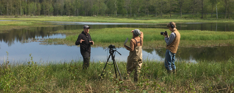 Tre personer står framför en våtmark med öppna vattenspeglar. Skog i bakgrunden. Två av personerna har varsin kamera och filmar den tredje personen. 