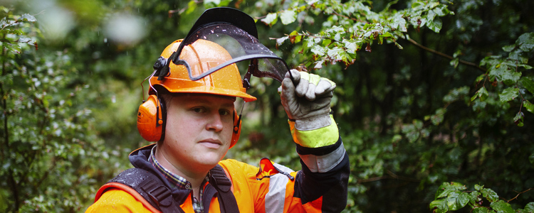 En manlig skogsarbetare klädd i orange arbetskläder står i en tät lövskog. Han lyfter på hjälmvisiret  och ser glad ut.