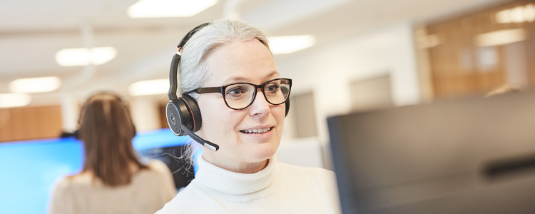 Kvinna med headset står vid ett höj- och sänkbart skrivbord och arbetar vid datorn på ett kontor, i ett kontorslandskap. Tittar koncentrerat in i skärmen. Foto. Foto: Sara Landstedt