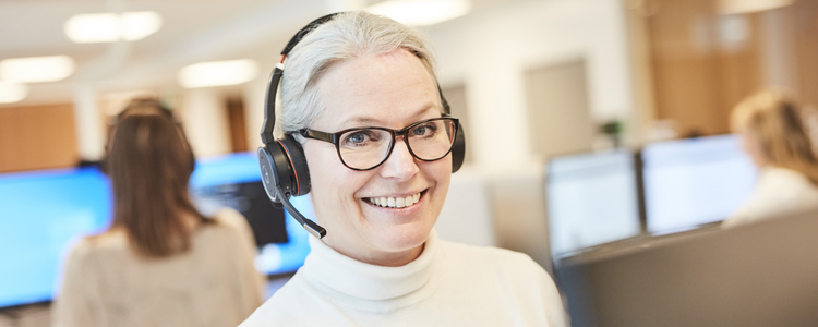 Kvinna med headset står vid ett höj- och sänkbart skrivbord och arbetar vid datorn på ett kontor, i ett kontorslandskap. Tittar glatt in i kameran. Foto. Foto: Sara Landstedt