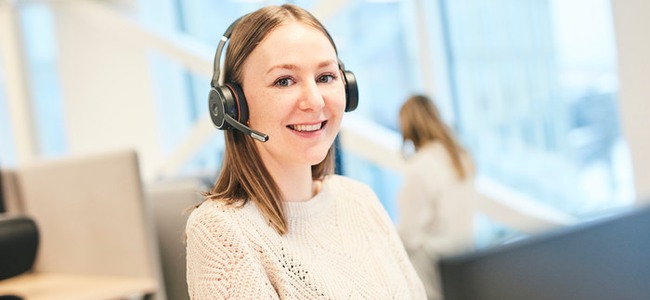Kvinna med headset står vid ett höj- och sänkbart skrivbord och arbetar vid datorn på ett kontor, i ett kontorslandskap. Tittar in i kameran och ler. Foto.